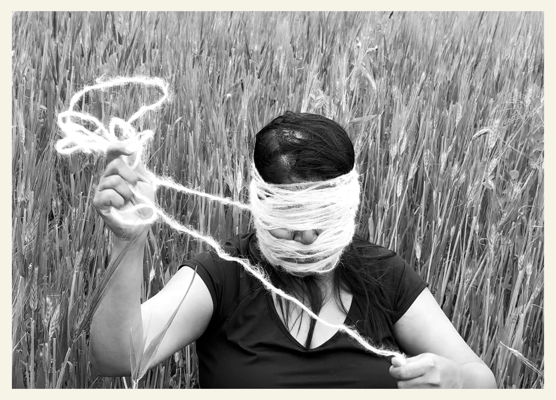 A figure's head wrapped in thread, held in a wheat field, in black and white